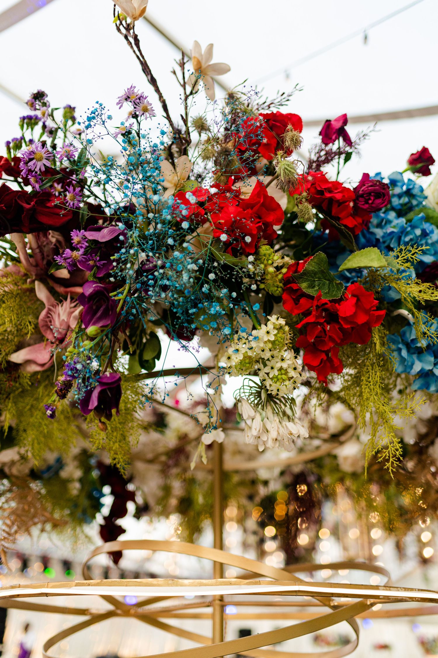 Lush floral chandelier with red, blue, and purple blooms on a gold frame under a white tent.