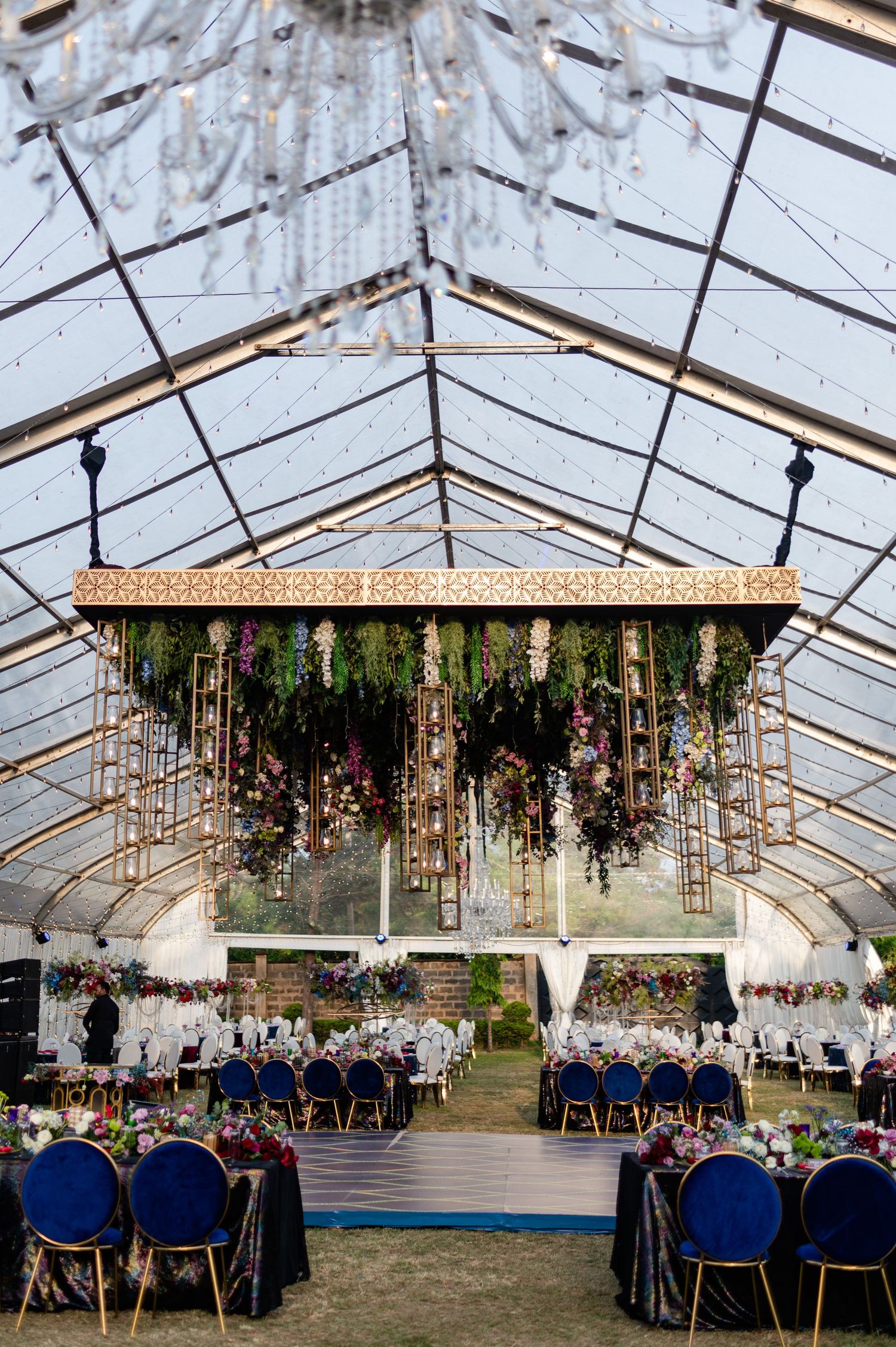Elegant wedding reception inside a clear tent with hanging floral installations, navy blue chairs, and crystal chandeliers.