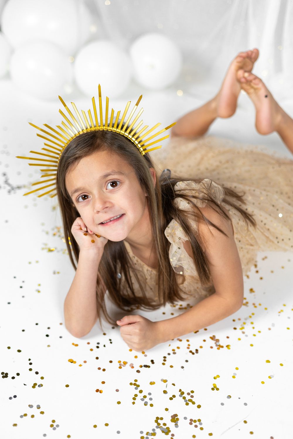Child in sparkly dress lying on floor with gold sunburst crown prop amidst glitter confetti.