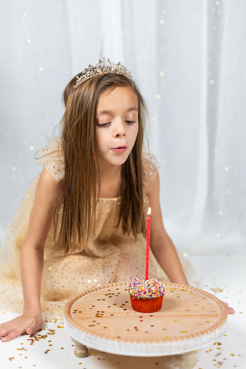 Child wearing tiara leaning over cupcake with lit candle on decorative white plate.