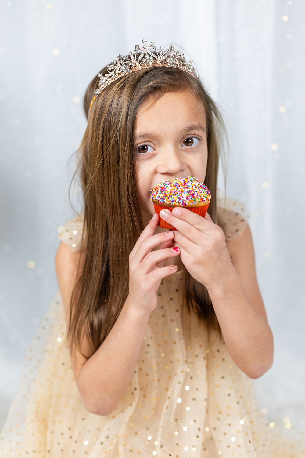 Child in sparkly dress wearing crown while eating colorful sprinkled cupcake.