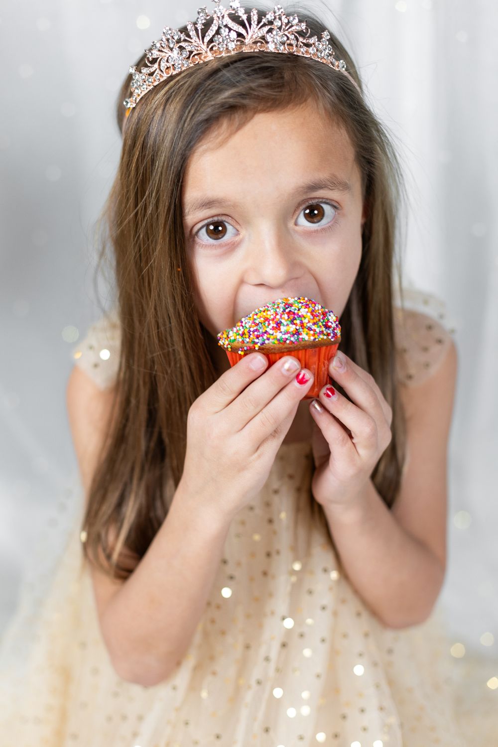 Child in tiara taking bite of sprinkle-covered cupcake against light background.