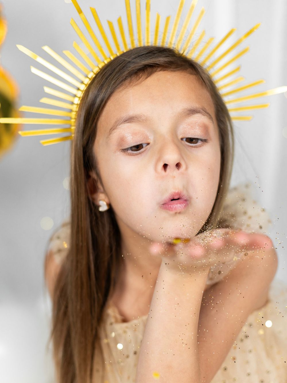 Child with golden sunburst crown blowing glitter from palm against white background.