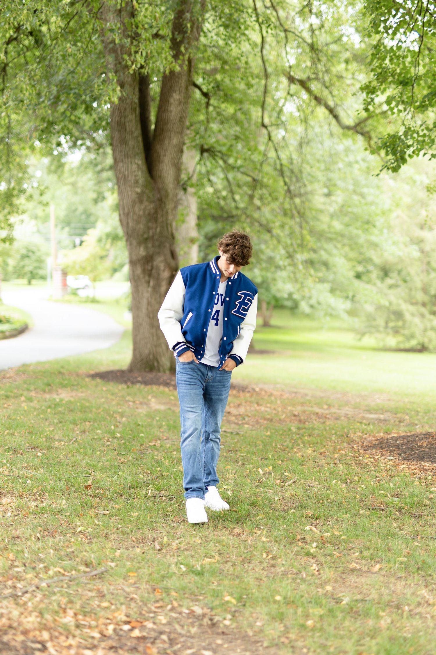 Person in denim and varsity jacket walking through a lush green park setting.