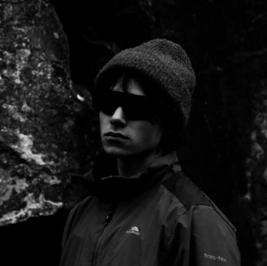 A person in a beanie and sunglasses poses against dark rocks in moody black and white.
