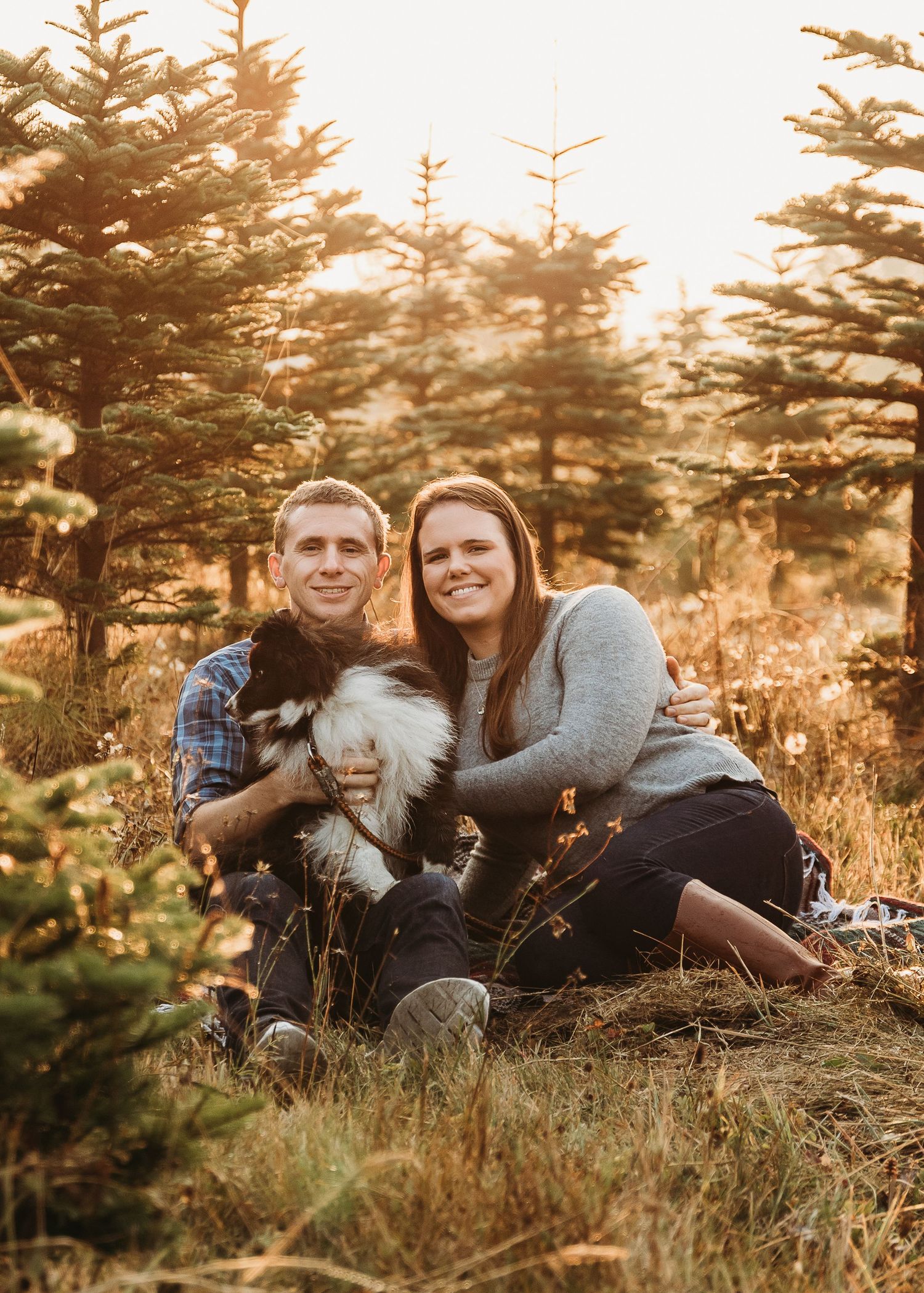 A couple sits with their fluffy white dog among evergreen trees during golden hour at sunset.