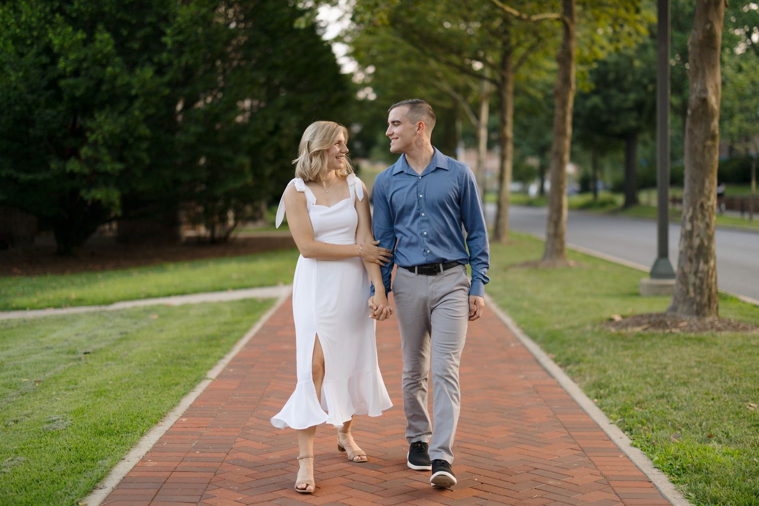 Couple walking together on a red brick pathway through a lush green park on a sunny day.