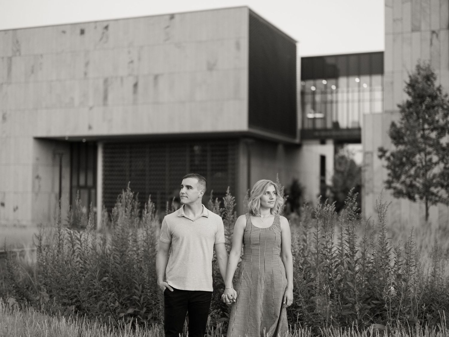 A couple stands together in front of a modern building surrounded by tall wild grasses in black and white.