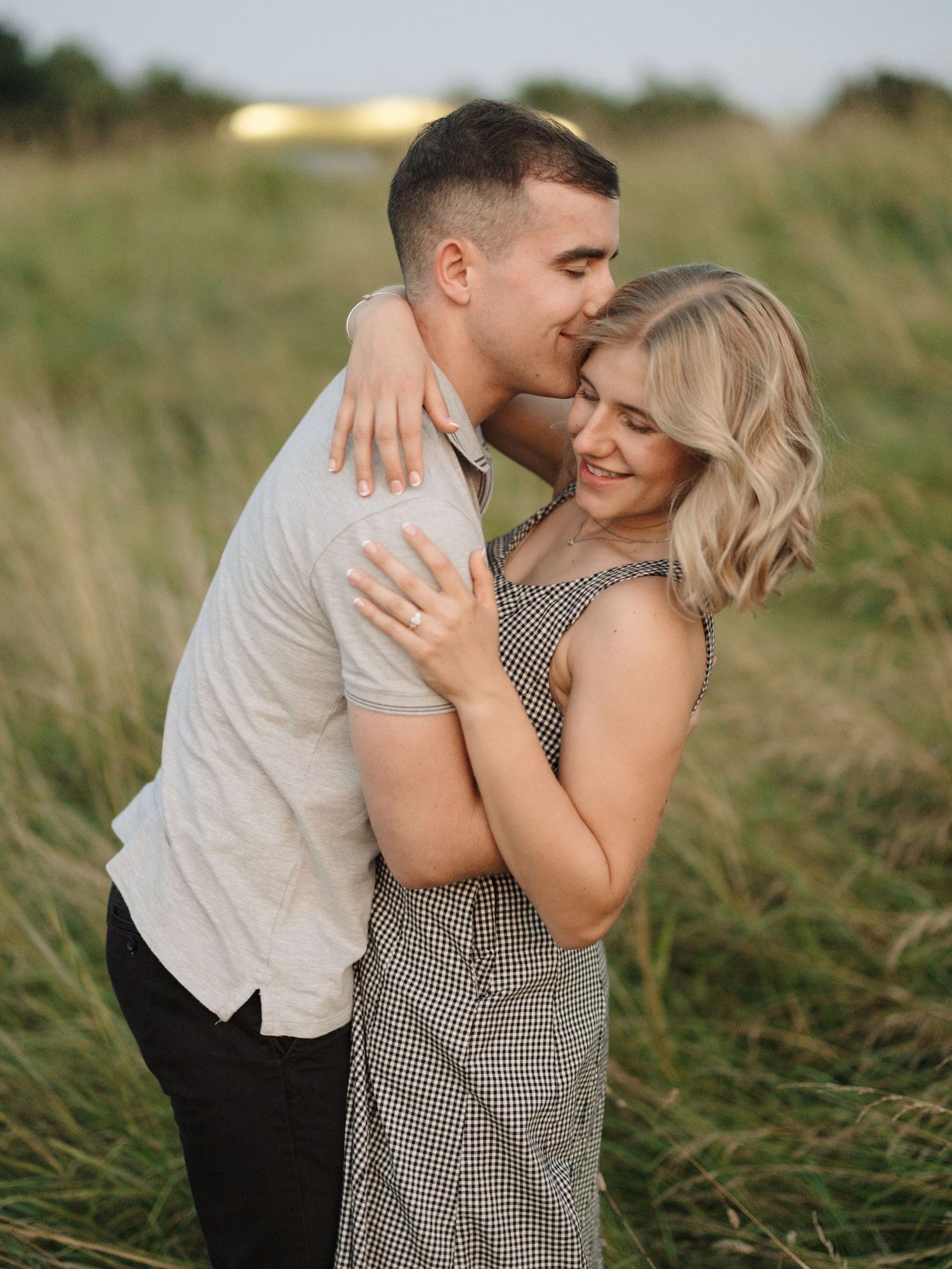 A romantic couple embraces in a field of tall grass during golden hour, with her wearing a checkered dress.