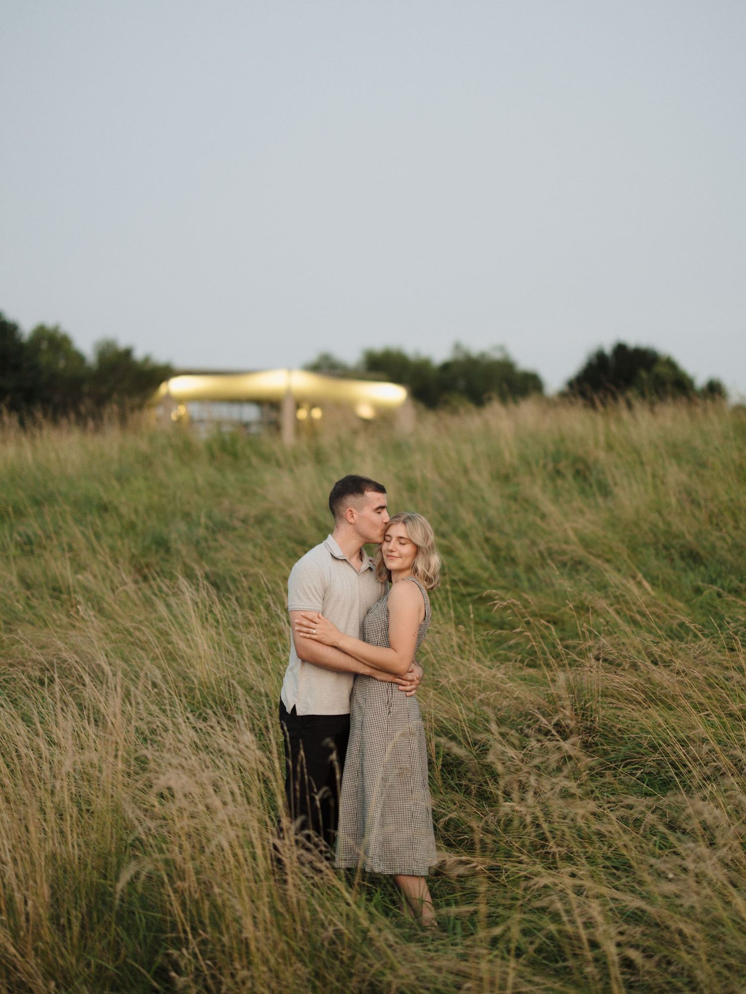 A loving couple shares a tender kiss while standing in a windswept meadow on a sunny afternoon.