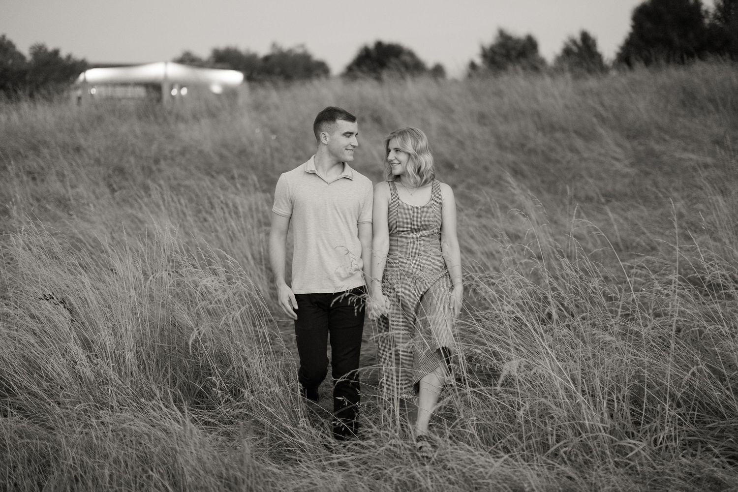A romantic black and white photo of a couple walking together through tall grass in a meadow.
