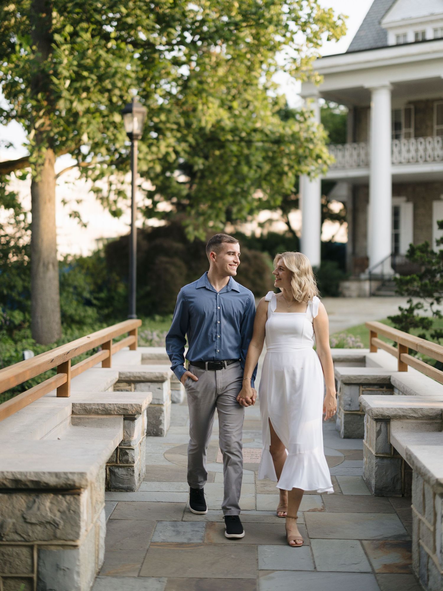 Couple enjoying a romantic walk together on a stone patio walkway during a sunny garden engagement session.