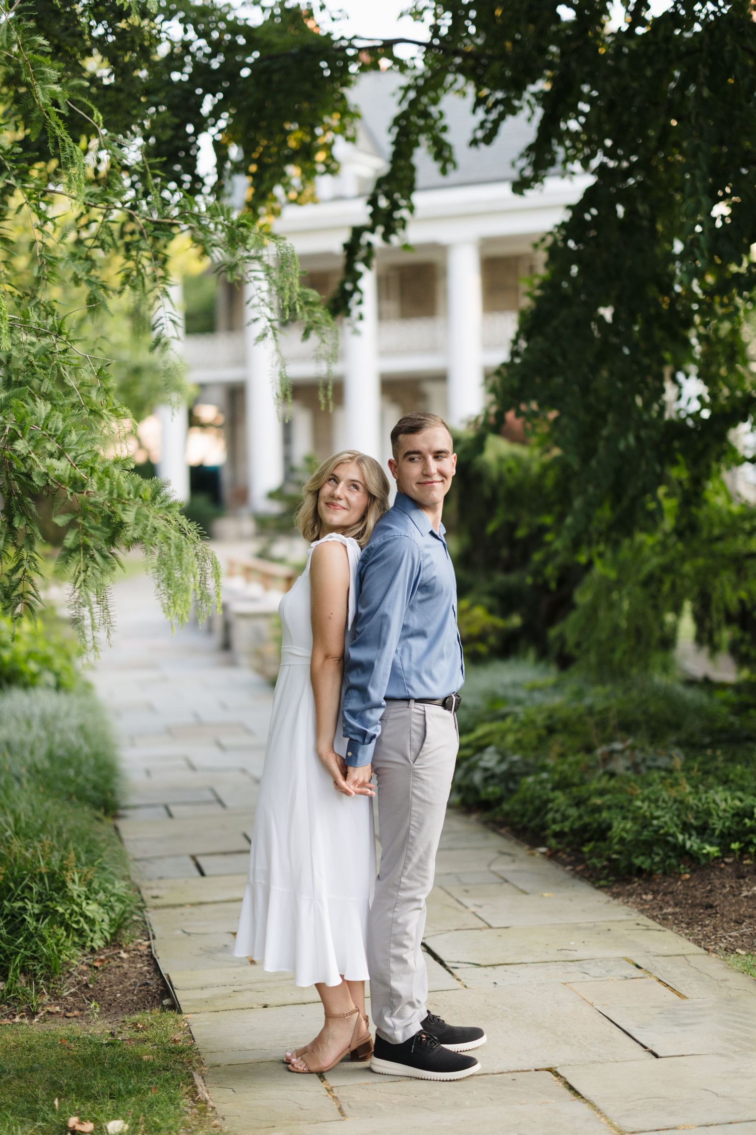 Couple poses back-to-back on stone path with white columned building and lush greenery in elegant garden setting.