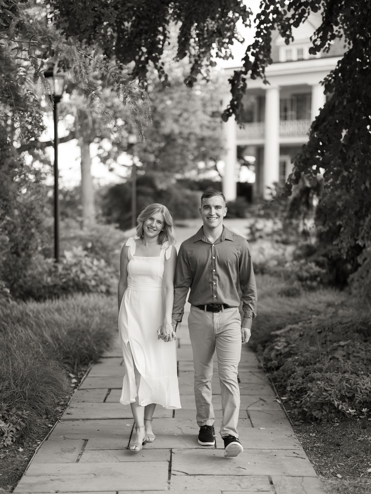 A couple walks hand in hand along a stone path with a stately white building visible in the background.