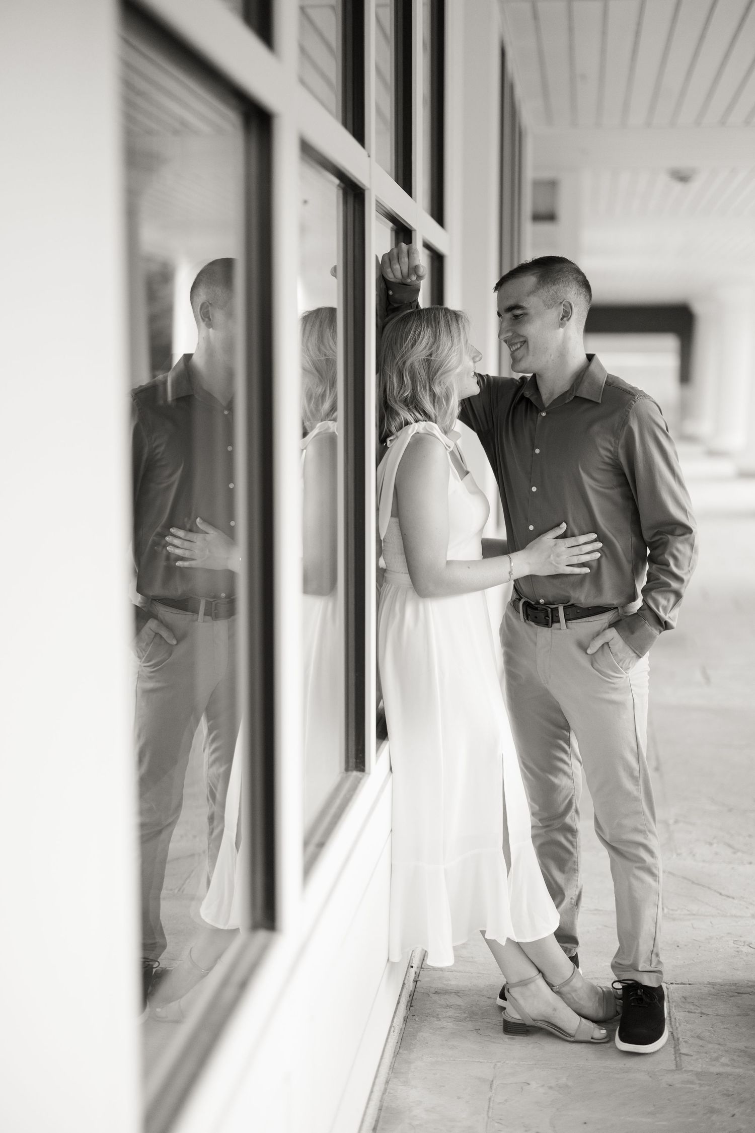 A romantic black and white portrait of a couple standing by large windows and sharing an intimate moment together.