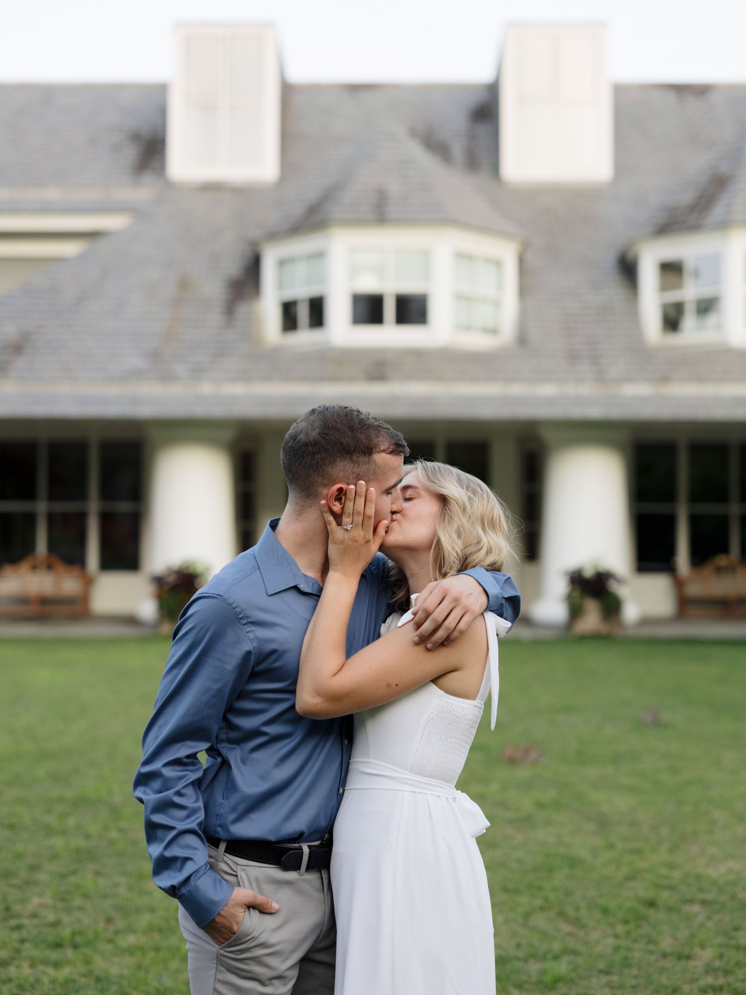 A couple in blue and white attire share a romantic kiss on a grassy lawn near a white building.