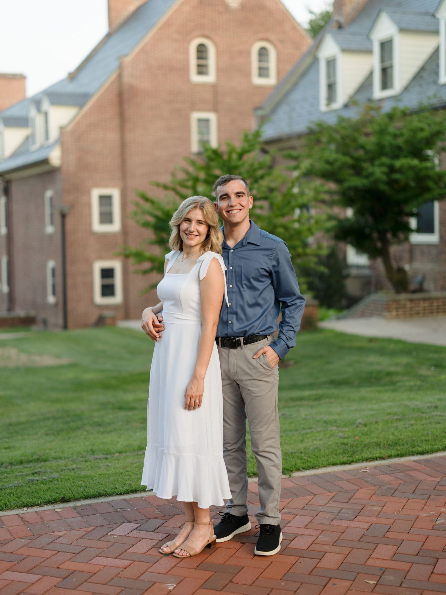 A couple poses progressively closer together on a brick pathway with brick buildings and green lawn in the background.