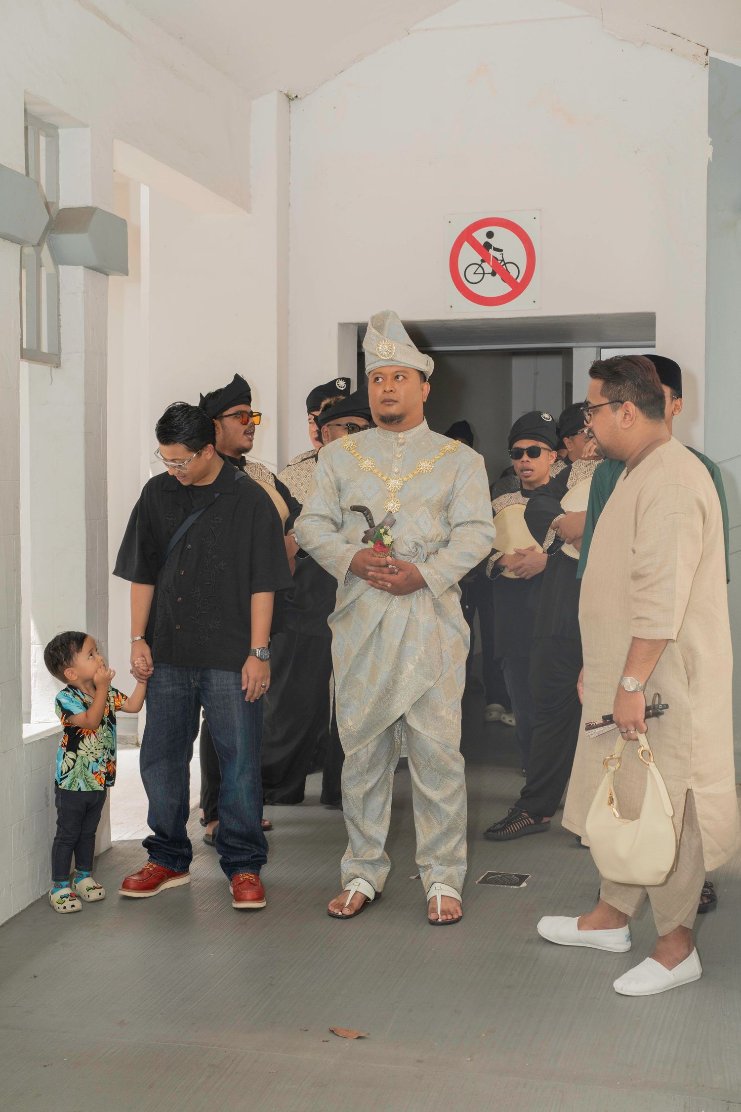 A group of people stand in a hallway beneath a no cycling sign, with one person wearing all white attire.