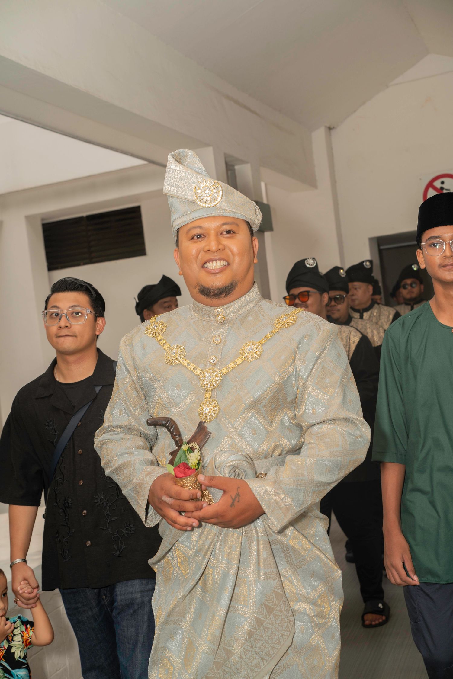 A groom in traditional Malay wedding attire with silver songkok and gold necklace poses during the ceremony.
