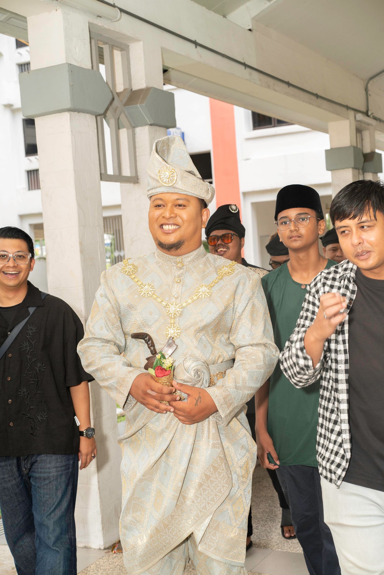 A group of people standing together at an event with one wearing traditional Malay ceremonial attire in cream color.