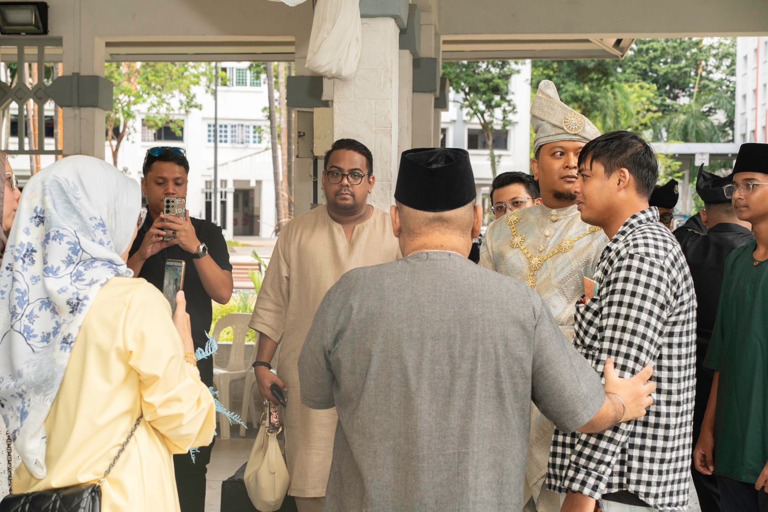 A group of people gather and interact in a covered outdoor area with sunlight visible in the background.