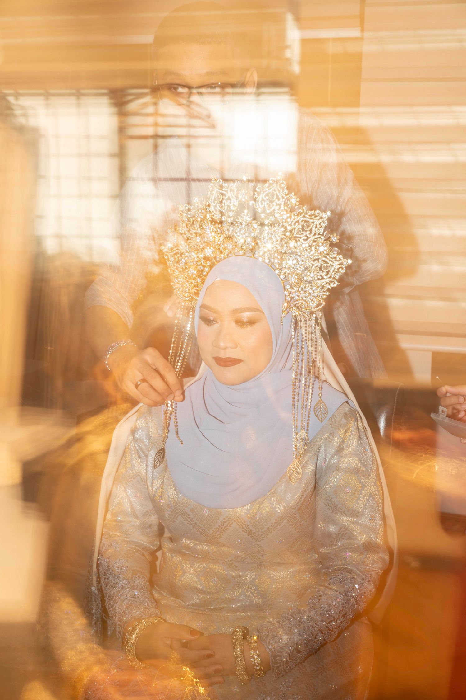 A bride in traditional Malaysian wedding attire with a blue hijab and ornate gold crown poses in soft warm lighting.