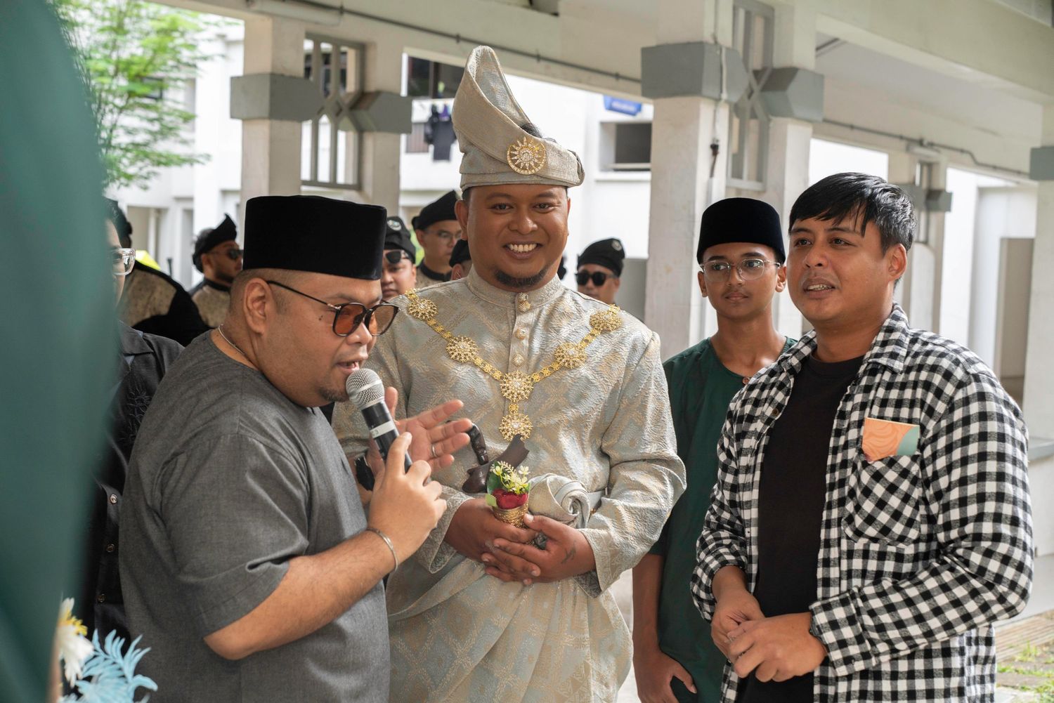 Several people gathered indoors, with one wearing traditional Malay wedding attire and others in casual clothing examining something.