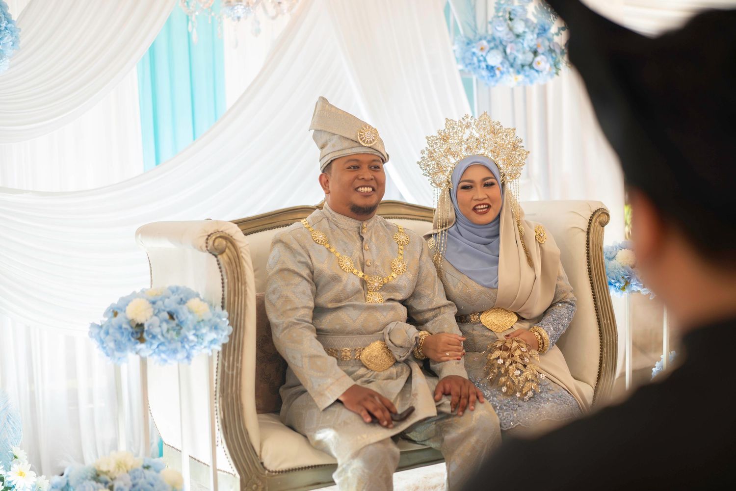 A wedding couple in traditional Malay attire sitting together on an elegant cream-colored sofa with blue and white decor.