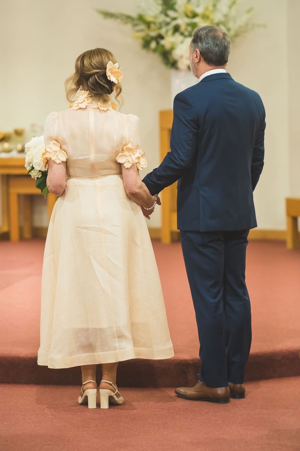 Couple stands at altar in wedding attire with bride wearing tea-length cream dress and groom in navy suit.