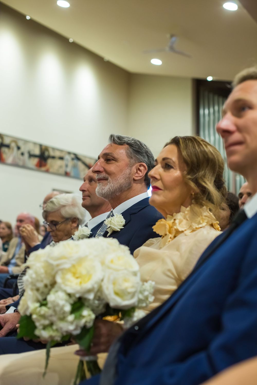 Wedding guests and couple sitting in the front row during a ceremony with white floral bouquet in view.
