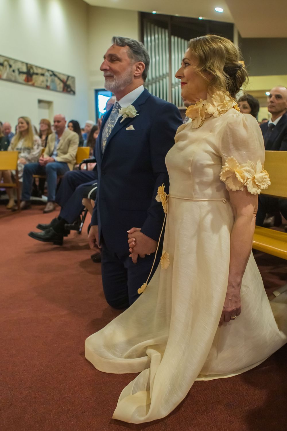 A couple stands together during their wedding ceremony wearing traditional formal attire in a church with guests seated behind.