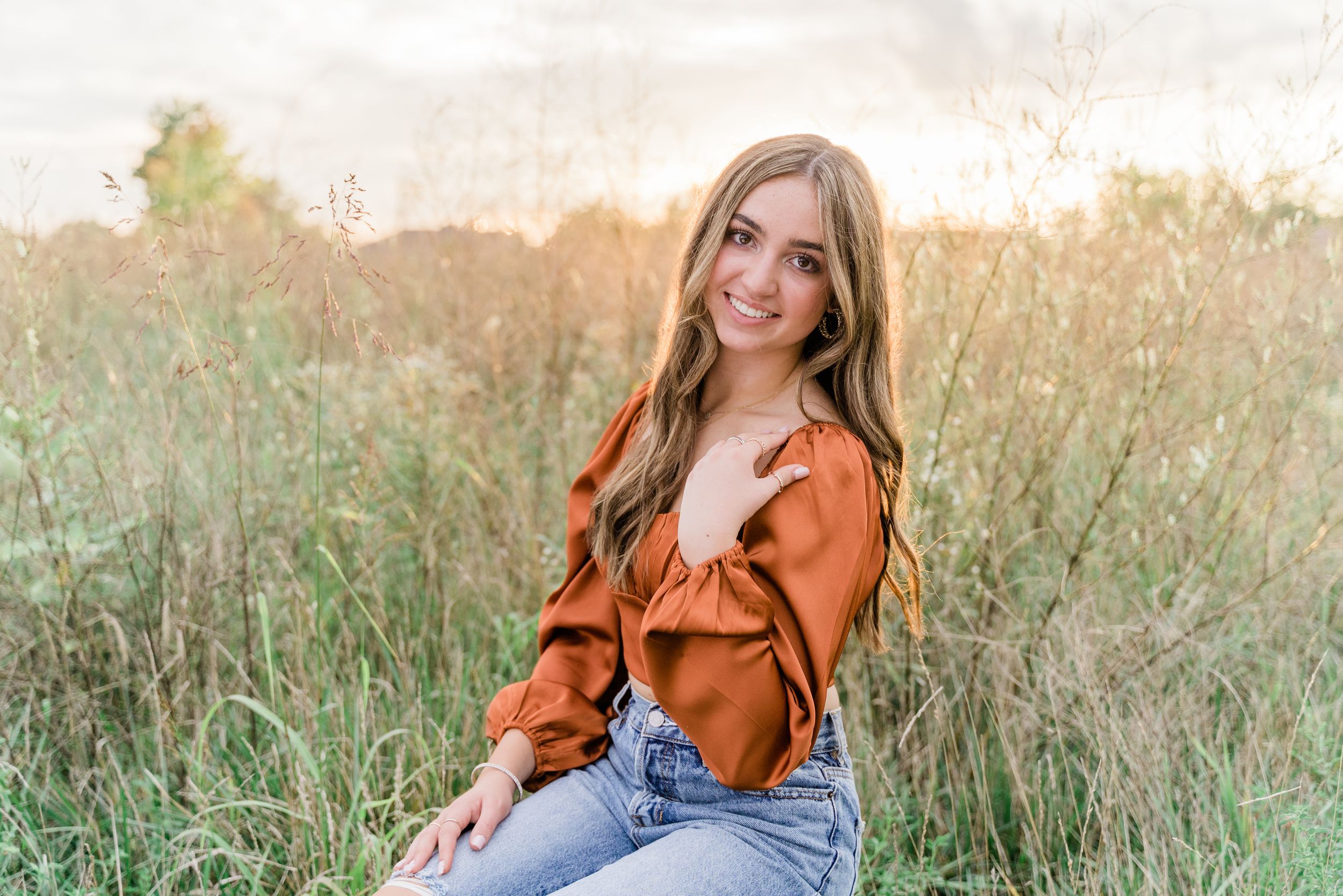 Woman in denim jeans and orange sweater sits casually in tall grass field during golden hour.