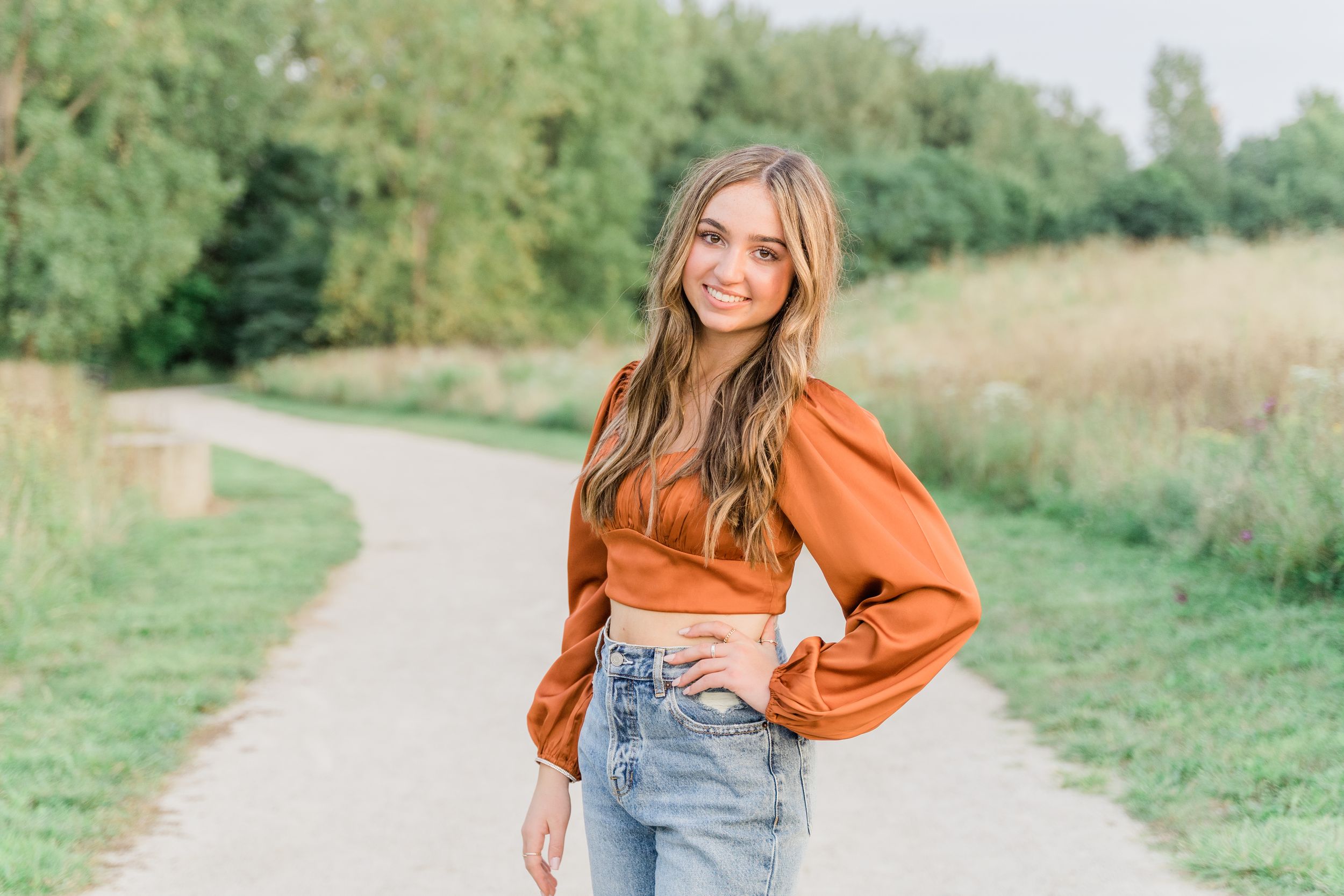 Woman in bright orange sweater and jeans poses outdoors on a grassy nature path.