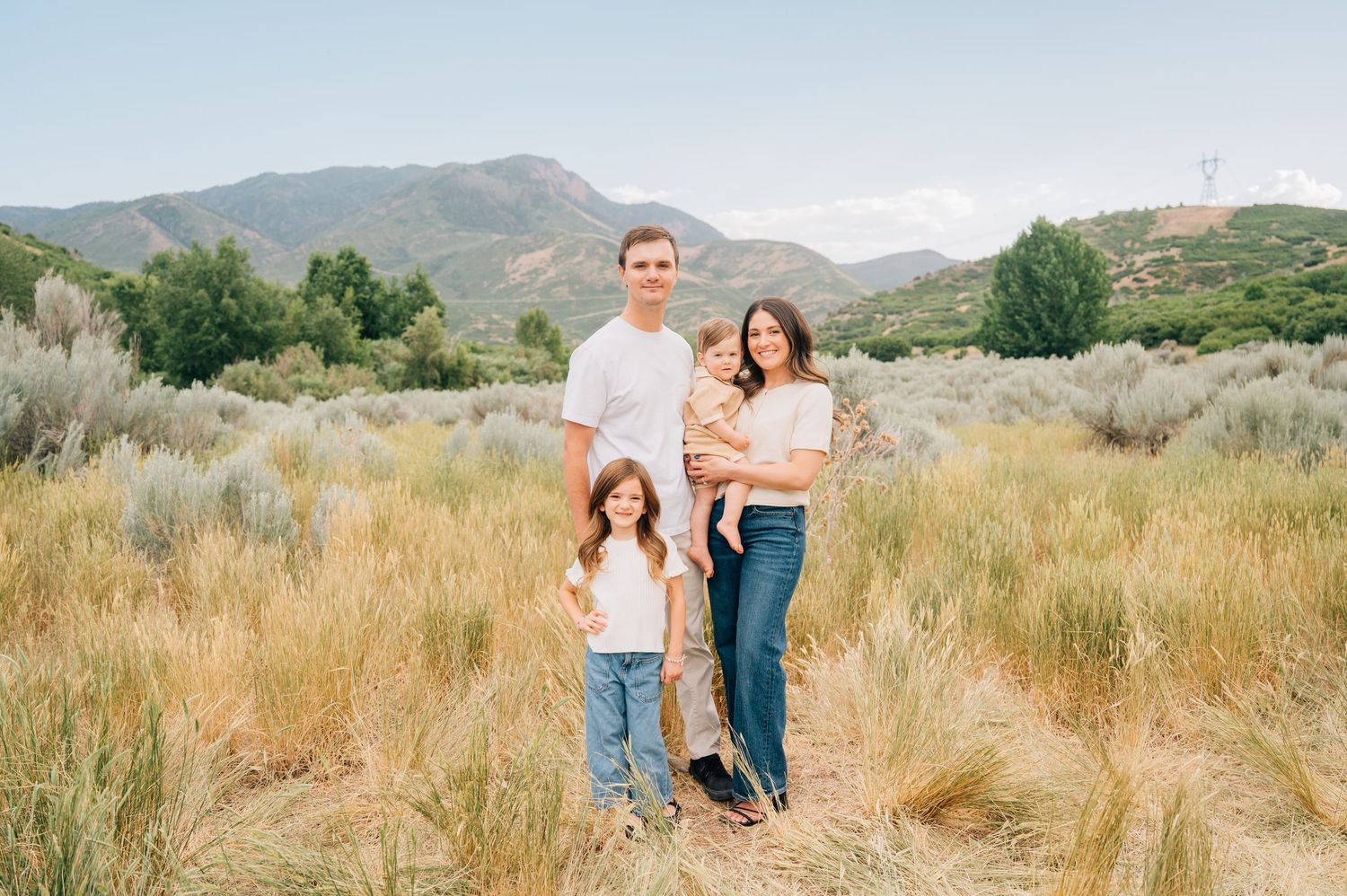 A family poses together in a field of tall grass with mountains and blue sky in the background.
