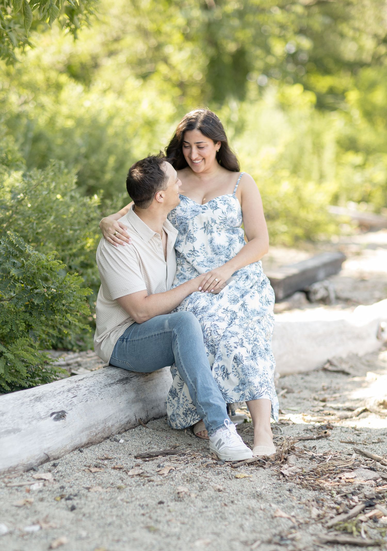A couple sits together on a rocky beach path surrounded by lush greenery on a sunny summer day.