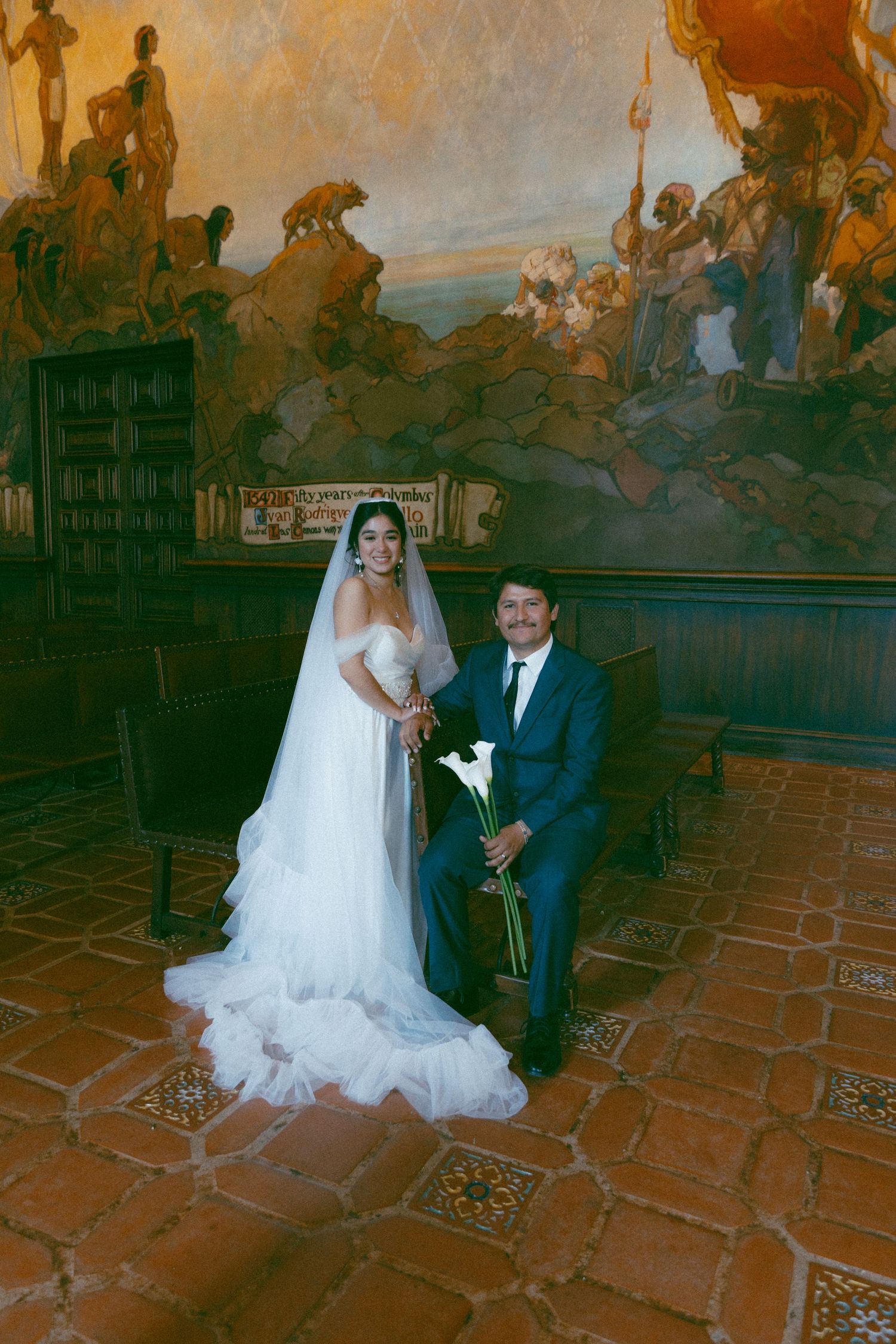 Newlyweds pose for a wedding portrait in an ornate chapel with historic murals and terracotta flooring
