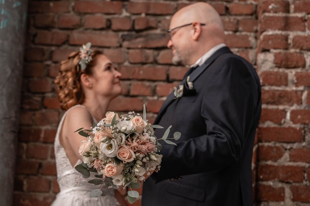 A romantic wedding moment between a couple sharing a tender look against a rustic brick wall with a blush rose bouquet.