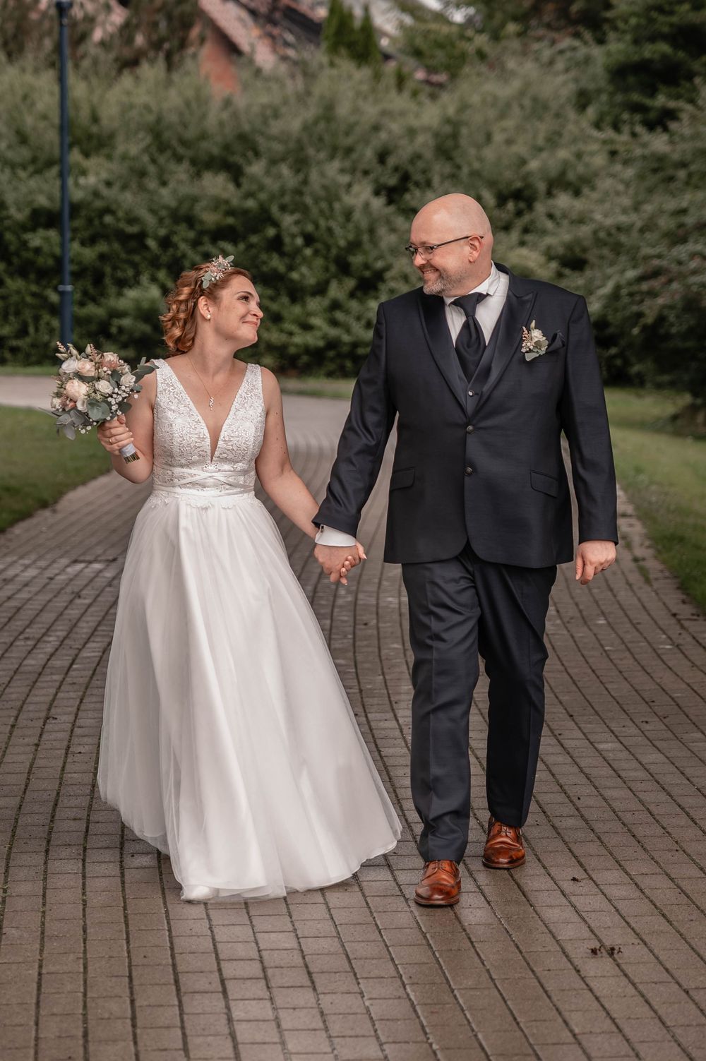 A bride in a white v-neck gown and groom in dark suit walk hand-in-hand along a brick pathway while sharing a happy moment.