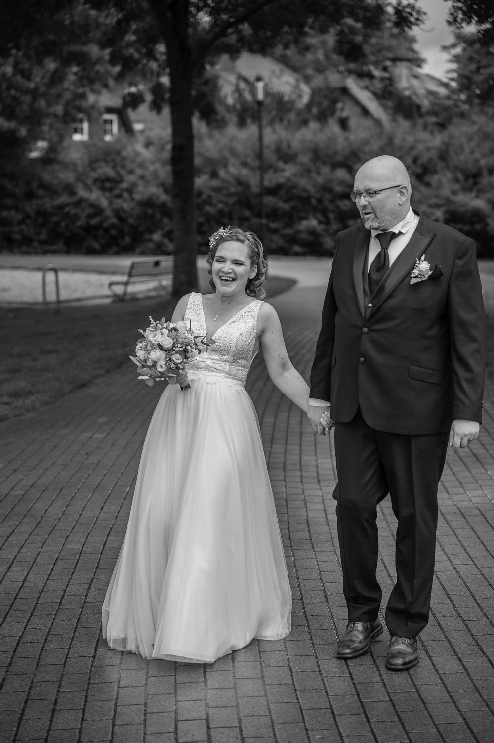 A flower girl in a white dress carries a bouquet while walking next to a person in a dark suit at an outdoor wedding.