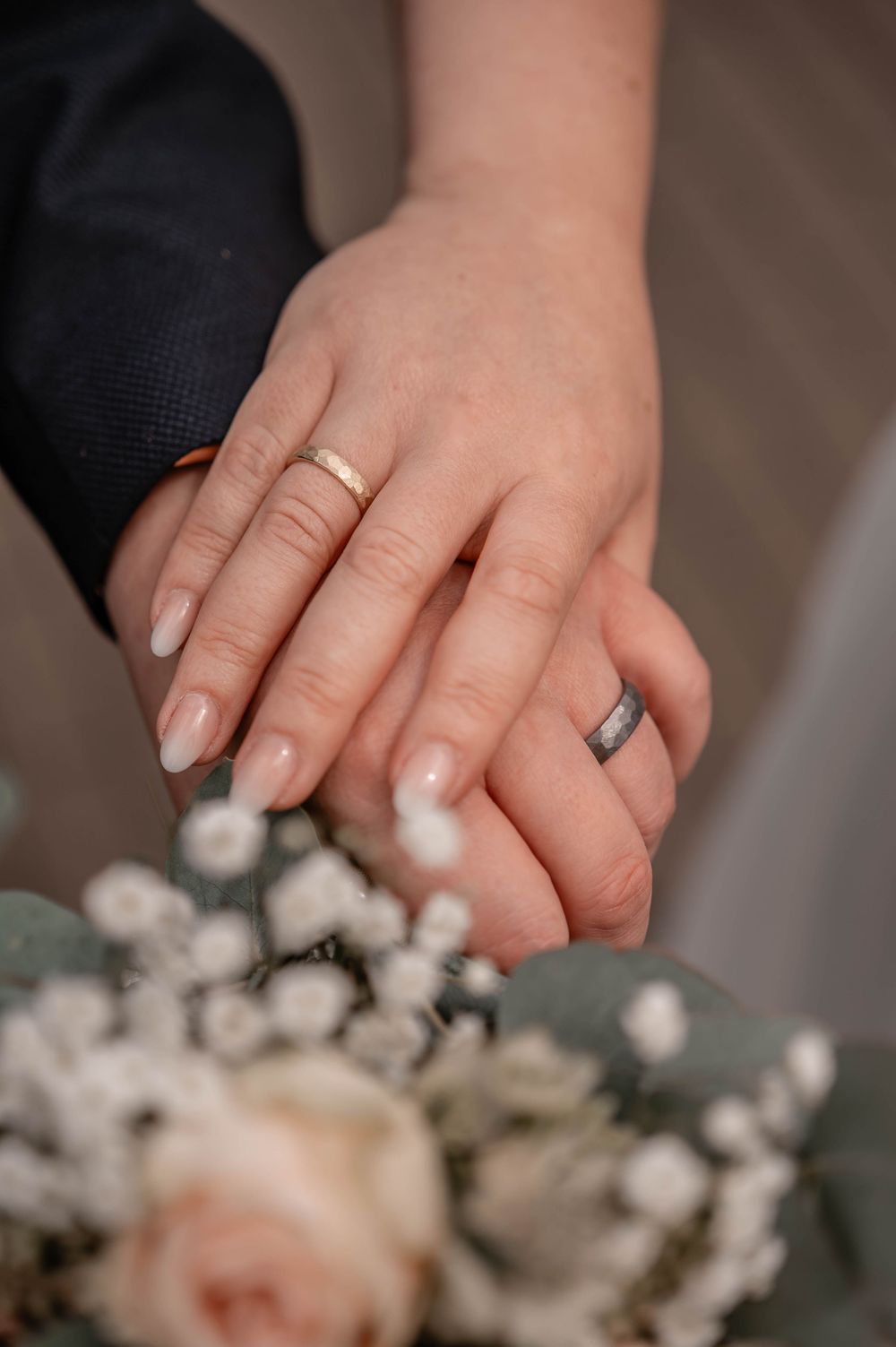 Close-up of newlyweds' hands with wedding rings resting above a soft pink and white floral bouquet.