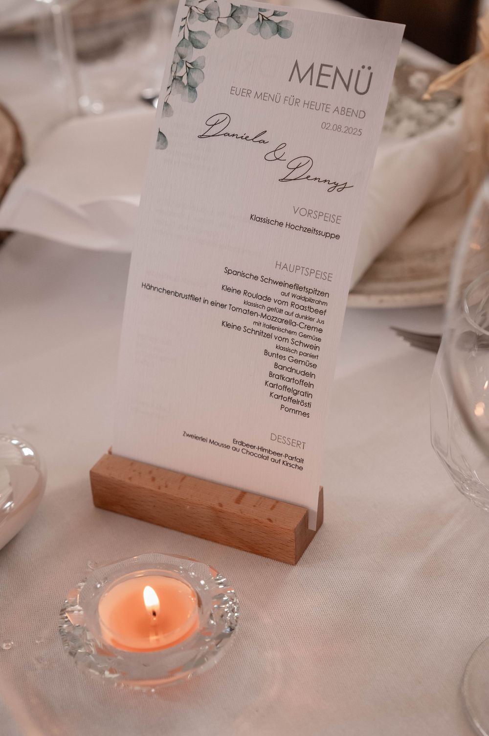 A wedding menu card displayed in a wooden holder on a white tablecloth with a lit peach-colored tealight candle nearby.