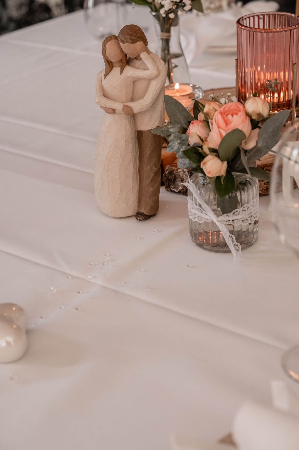 Wedding decor featuring a Willow Tree figurine, pink roses in a lace-wrapped vase, and a coral glass candleholder on white linen.