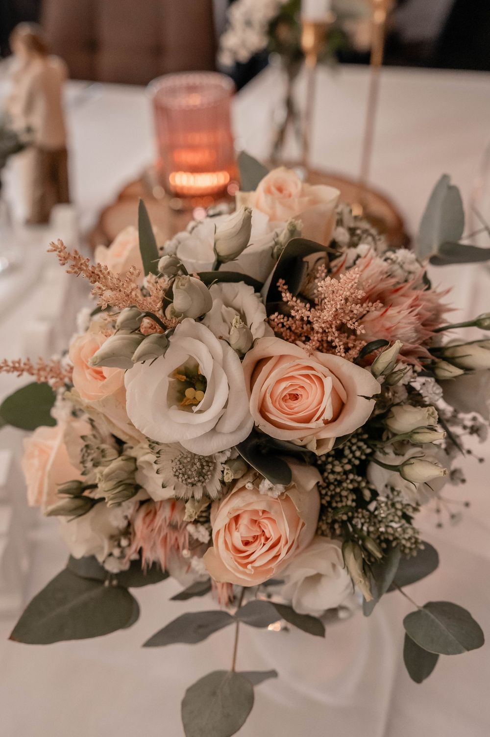 Romantic wedding centerpiece featuring peach roses and white flowers with eucalyptus leaves and a glowing candle in soft lighting.