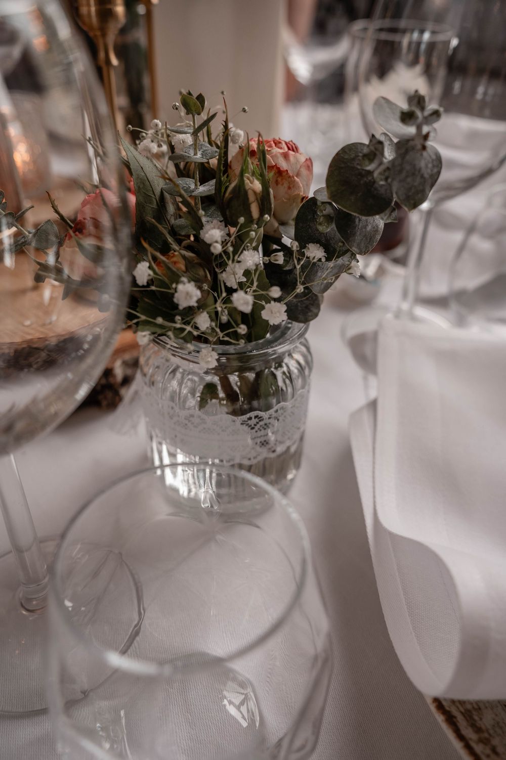 A delicate floral arrangement with roses and eucalyptus in a vintage cut glass vase sits among wine glasses on a white table.