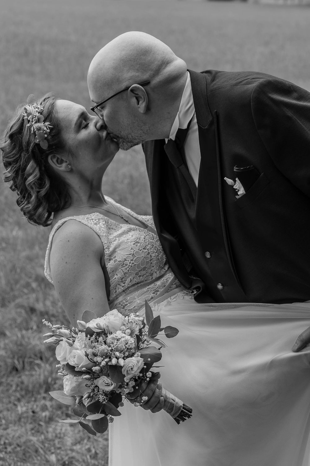 A black and white wedding photograph capturing a romantic kiss with a floral bouquet in hand and elegant lace dress details.