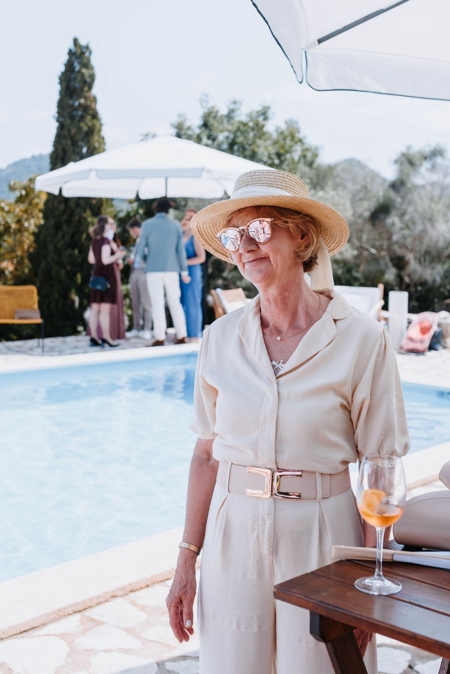 Die Mama in einem cremefarbenen Overall und einem Sonnenhut genießt an einem sonnigen Sommertag den Anblick ihrer Töchter am Pool.