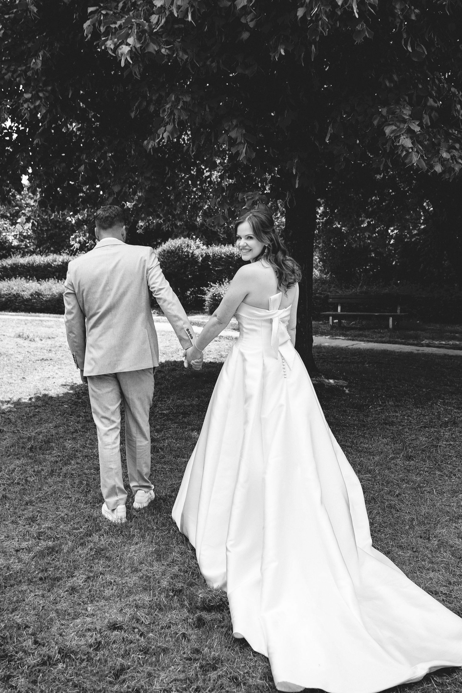 A black and white wedding photo showing a couple walking hand in hand through a garden with a long flowing wedding gown train.