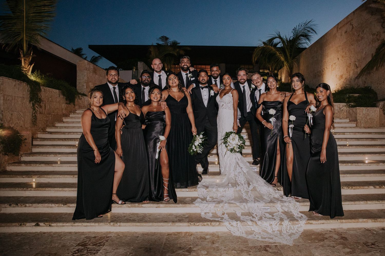 Wedding party in black attire poses on stone steps for a formal group photo.
