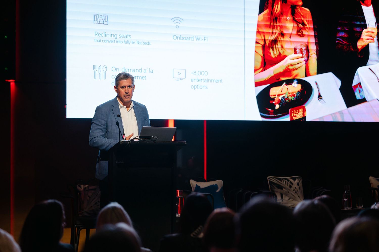 A business conference presentation shown across multiple screens with speakers and audience members in a dark auditorium.