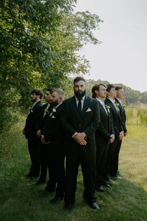 Group of groomsmen in dark suits gathered on grassy lawn.